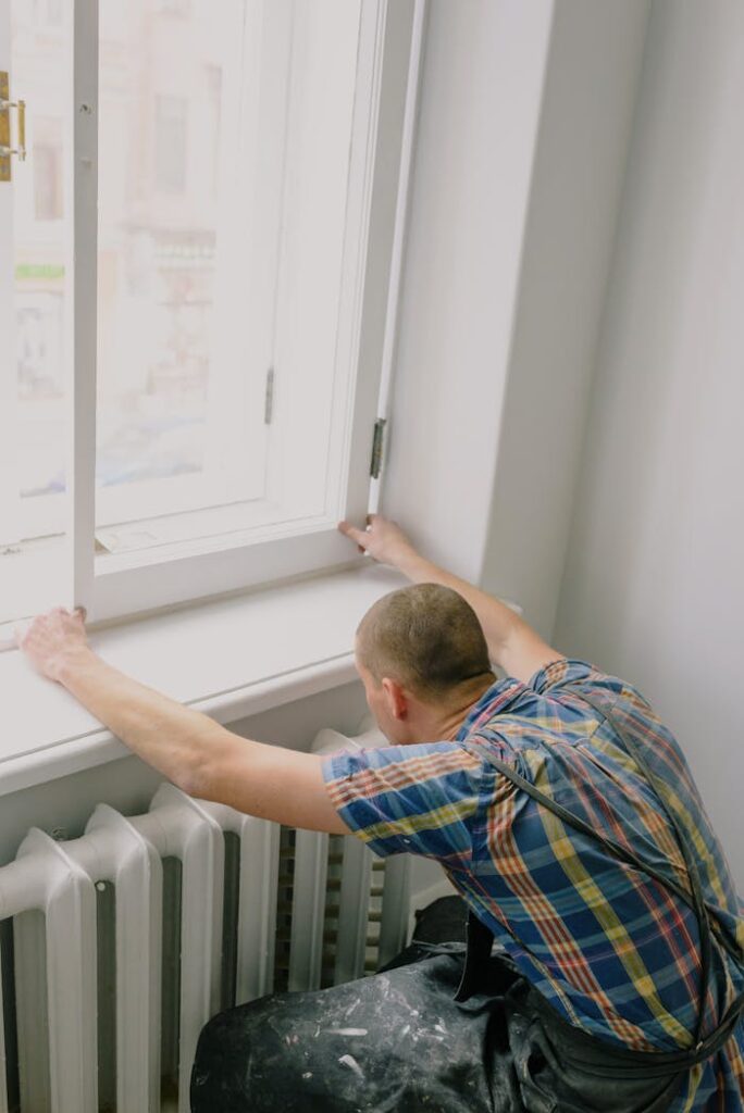 Back view of unrecognizable fitter in dirty apron installing window above radiator while doing home renovation